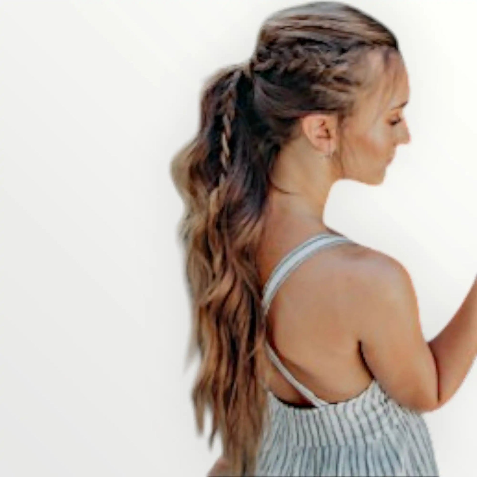 Een vrouw met lang, golvend bruin haar, gestyled in een halve opgestoken gevlochten paardenstaart, draag een gestreepte tanktop.